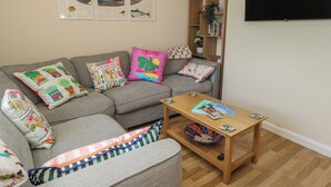 A living room with a grey sofa, colorful pillows, a wooden coffee table, and wall-mounted artwork. - Mallow Cottage (Swanage)