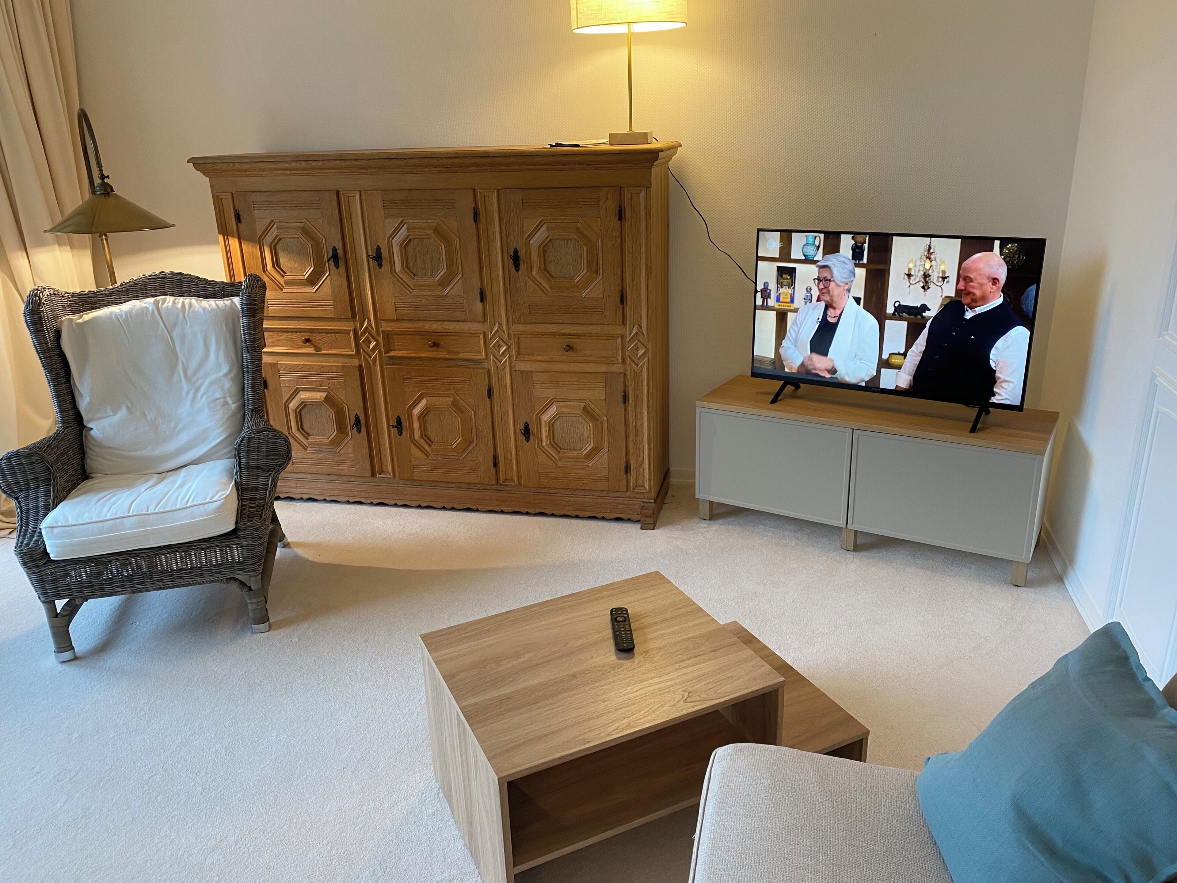 Uma sala de estar com uma poltrona de vime, uma mesa de centro de madeira, uma televisão sobre um móvel e um armário.