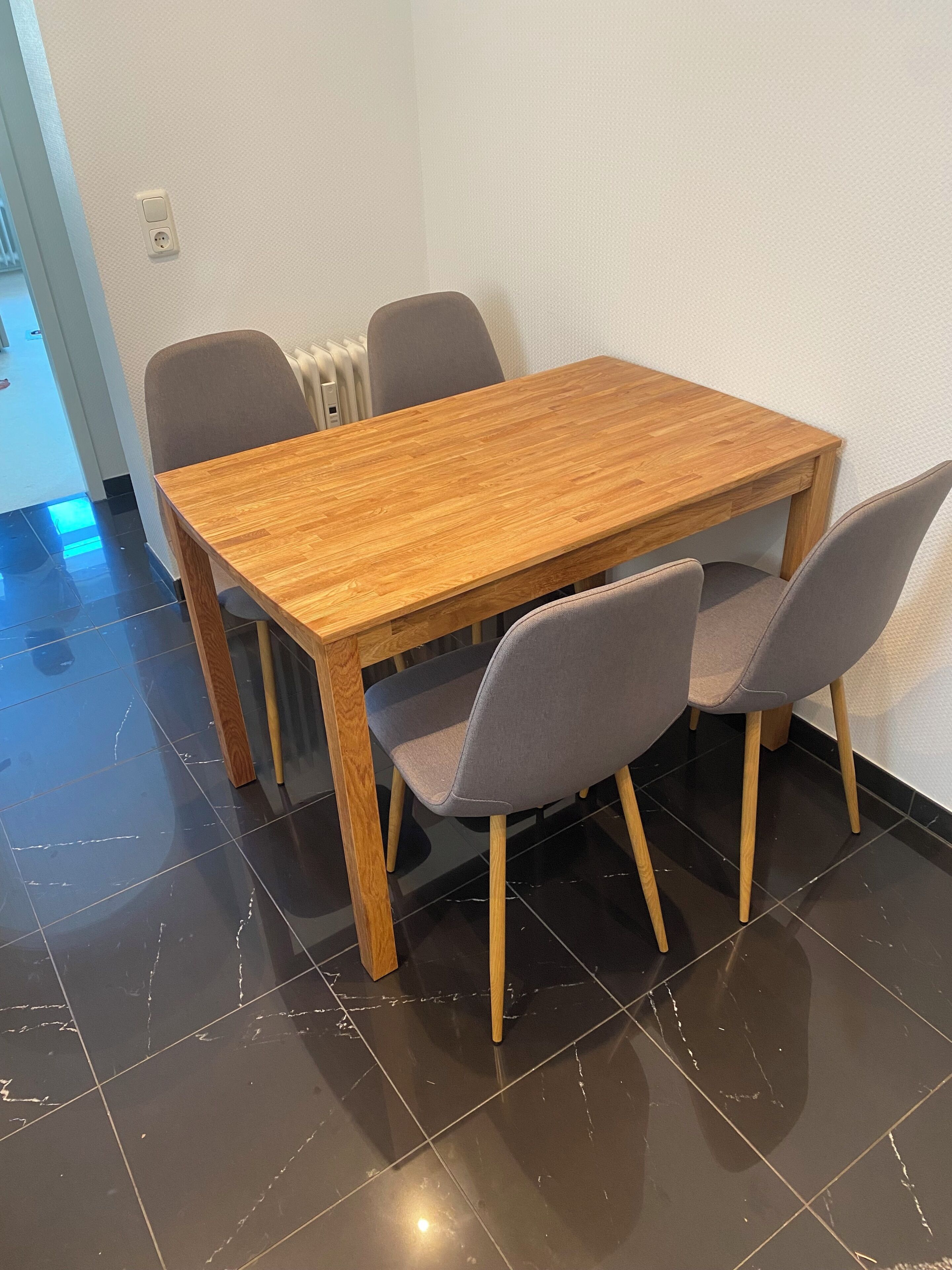 Una mesa de comedor de madera con cuatro sillas grises, una pared blanca y un radiador.