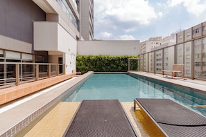Ein Schwimmbecken mit Liegestühlen, einer Hecke und modernen Gebäuden im Hintergrund.