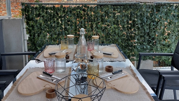 An outdoor dining area with a table set for two, surrounded by a green leafy wall, with a brick building in the background. - Maisonette with south-facing terrace & barbecue (Kapsweyer)
