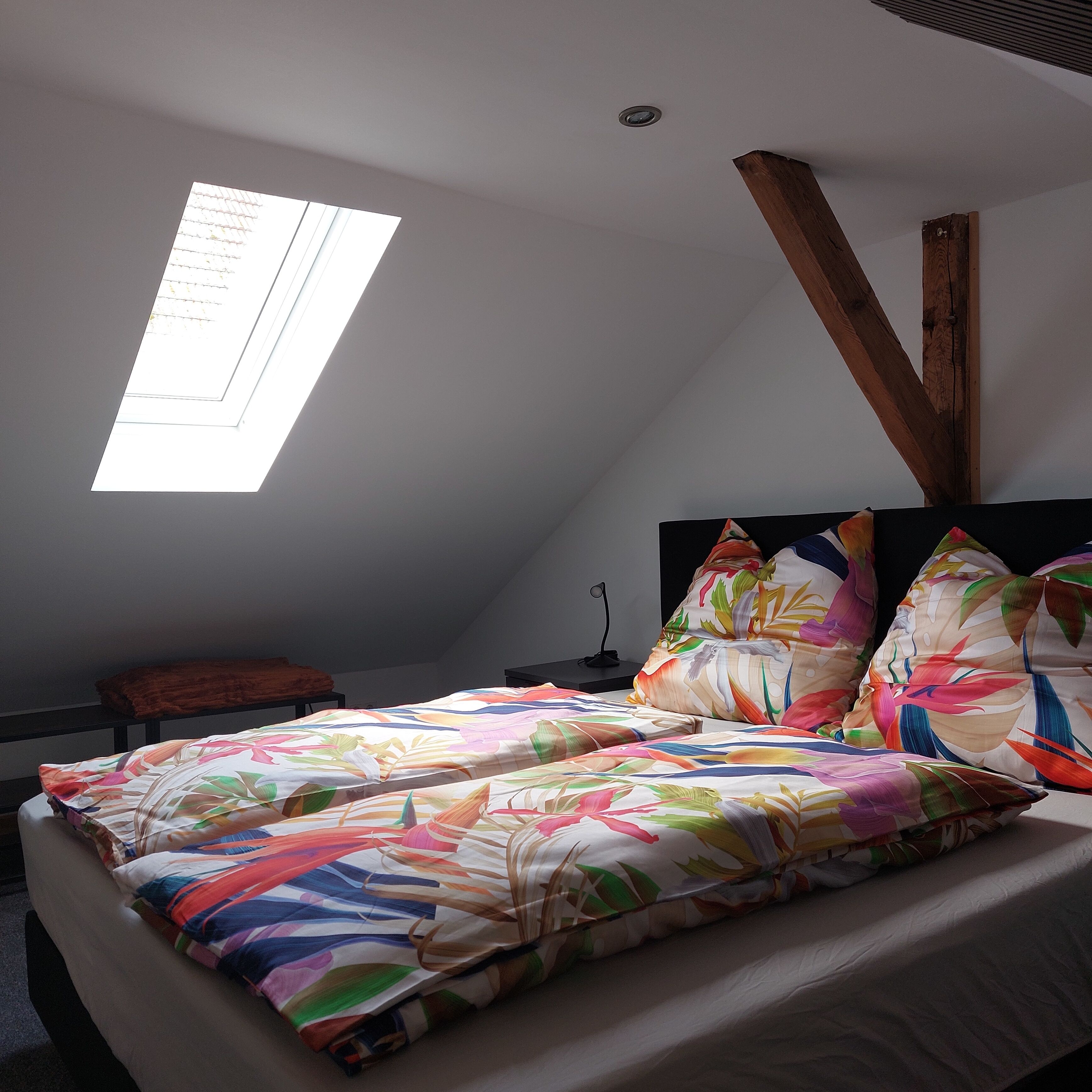 A bedroom with a sloped ceiling, a skylight, a bed with colorful floral bedding, a wooden headboard, and a small bedside table.