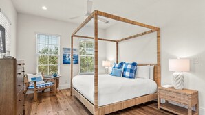 Een hemelbed met houten frame, een nachtkastje met lamp, een stoel met geruit kussen, en een raam met jaloezieën.