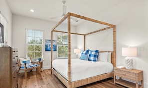 Ein Bett mit Baldachin und Holzrahmen, ein Nachttisch mit Lampe, ein Sessel mit kariertem Kissen und ein Fenster mit Jalousien.