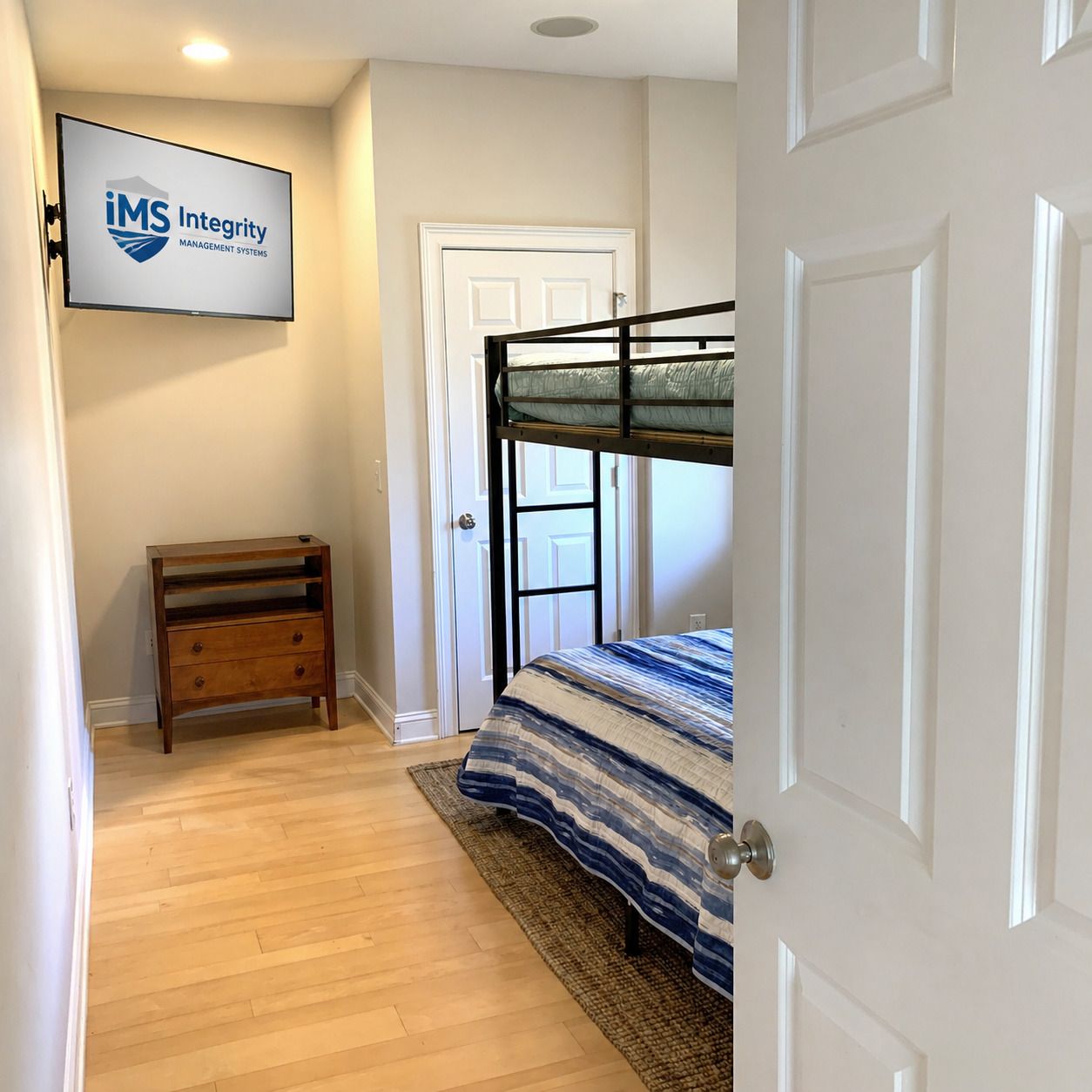 Un dormitorio con una cama litera, una mesita de noche de madera, un logotipo enmarcado en la pared y una puerta blanca.