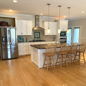A modern kitchen with white cabinets, a stainless steel refrigerator, a central island with bar stools, and pendant lighting. - Salt & Lime at Turner Tide (North Myrtle Beach)