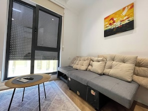 Ein Wohnzimmer mit grauem Sofa, einem kleinen runden Tisch und einem großen Fenster mit Jalousien.