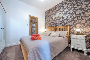 A bedroom with a bed, a wooden headboard, a nightstand with a lamp, a dresser, and a floral wallpaper. - Ground Floor 1-bed Apartment at Sea-cote (Blackpool)