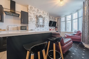 A modern kitchen with a flat-screen TV, a microwave, and a coffee maker. There are two bar stools and a red leather sofa. - Ground Floor 1-bed Apartment at Sea-cote (Blackpool)
