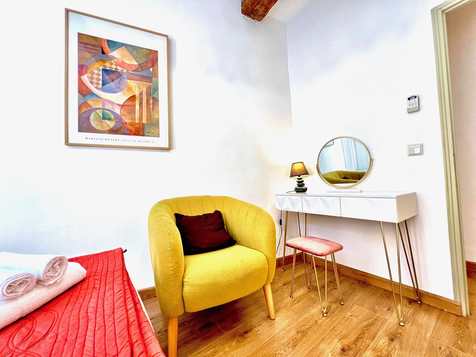 Una habitación con un sillón amarillo, un escritorio blanco con un espejo, una colcha roja y un cuadro abstracto enmarcado.
