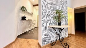 Een gang met een houten vloer, een witte muur met bladmotief, een klein tafeltje met een plant en een stoel.