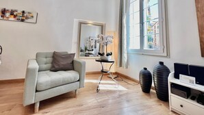 Een moderne woonkamer met een lichtgrijze bank, een grote spiegel, een raam met uitzicht op gebouwen en een zwarte speaker op een witte plank.