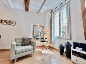 Een moderne woonkamer met een lichtgrijze bank, een grote spiegel, een raam met uitzicht op gebouwen en een zwarte speaker op een witte plank.