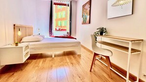 Een moderne slaapkamer met een bed, nachtkastje, bureau, stoel en een groot raam met uitzicht op gebouwen.