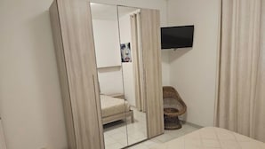 A bedroom with a bed, a wardrobe with a mirror, a television on the wall, and a wicker chair. - The villa is located 250 meters from the sandy beaches with crystal clear sea. (Baia Verde)