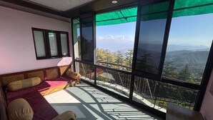 Una habitación con balcón, un sofá con cojines, una ventana con vistas a montañas y un ventilador de techo.