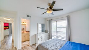 A bedroom with a bed, a ceiling fan, a window with blinds, and a bathroom with a vanity and mirror.