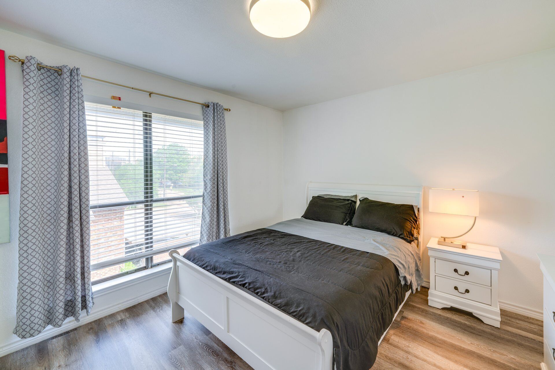 Een slaapkamer met een bed, een nachtkastje met een lamp en een raam met gordijnen.