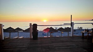 Eine Holzterrasse mit Blick auf einen Strand, Schilmdächer und einem Sonnenuntergang.