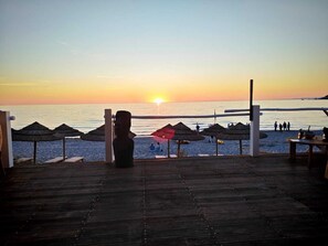 Eine Holzterrasse mit Blick auf einen Strand, Schilmdächer und einem Sonnenuntergang.