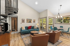 A modern living room with a blue sofa, brown armchairs, a coffee table, a TV, and a spiral staircase. - Cozy Mountain Condo - Pool, Sauna, & Hot Tub Access (Keystone)