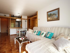 A living room with a sofa, a dining table, and chairs. There is a kitchen area with cabinets and a refrigerator. A painting is on the wall. - Apartment with AC and pool - 4 guests (Lloret de Mar)
