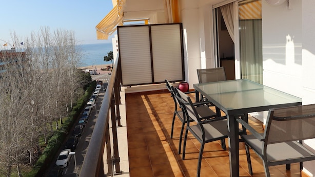 Un balcón con una mesa y sillas de vidrio, vista al mar y un edificio de fachada blanca.