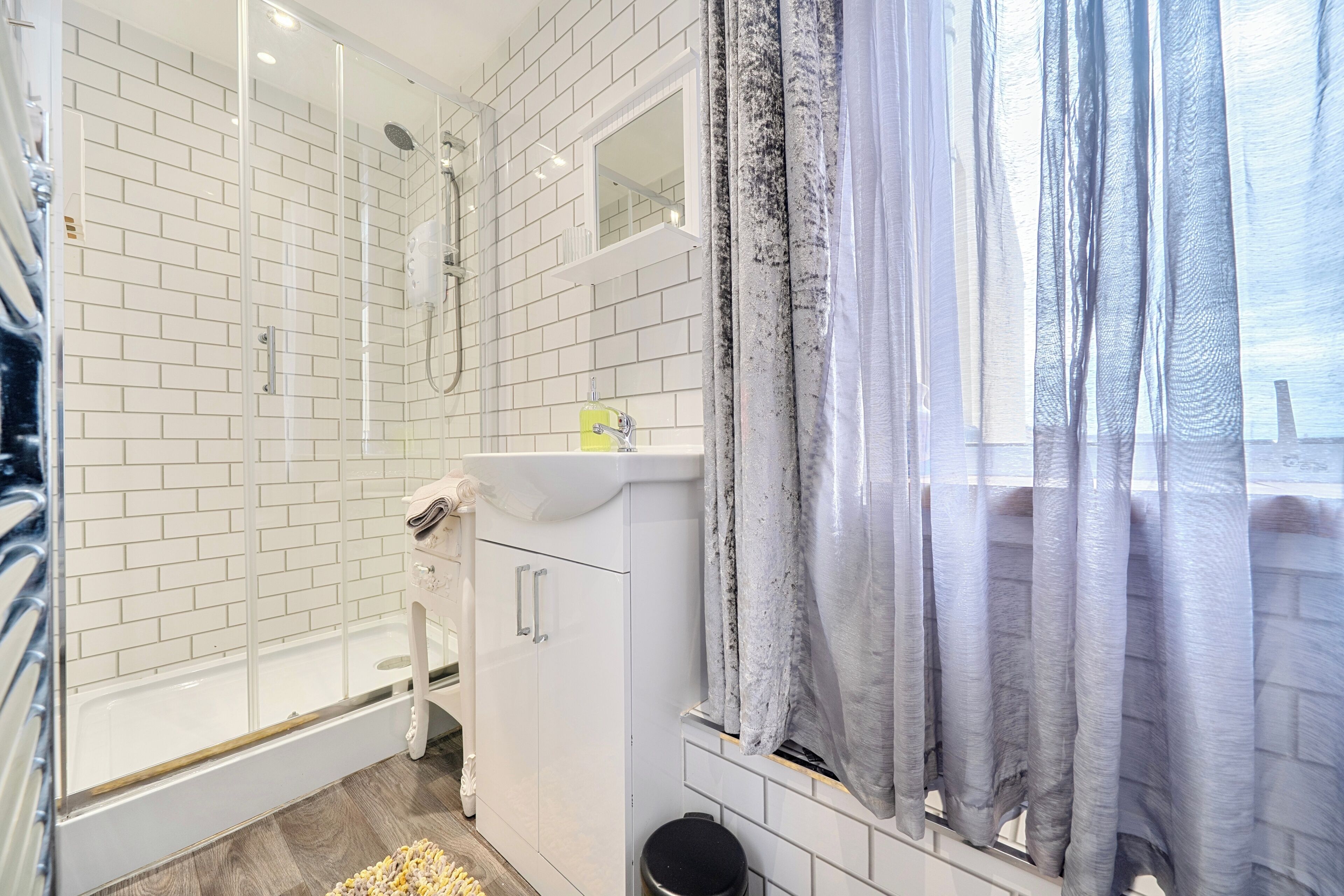 A bathroom with white tiled walls, a glass shower enclosure, a white sink, and a window with curtains.