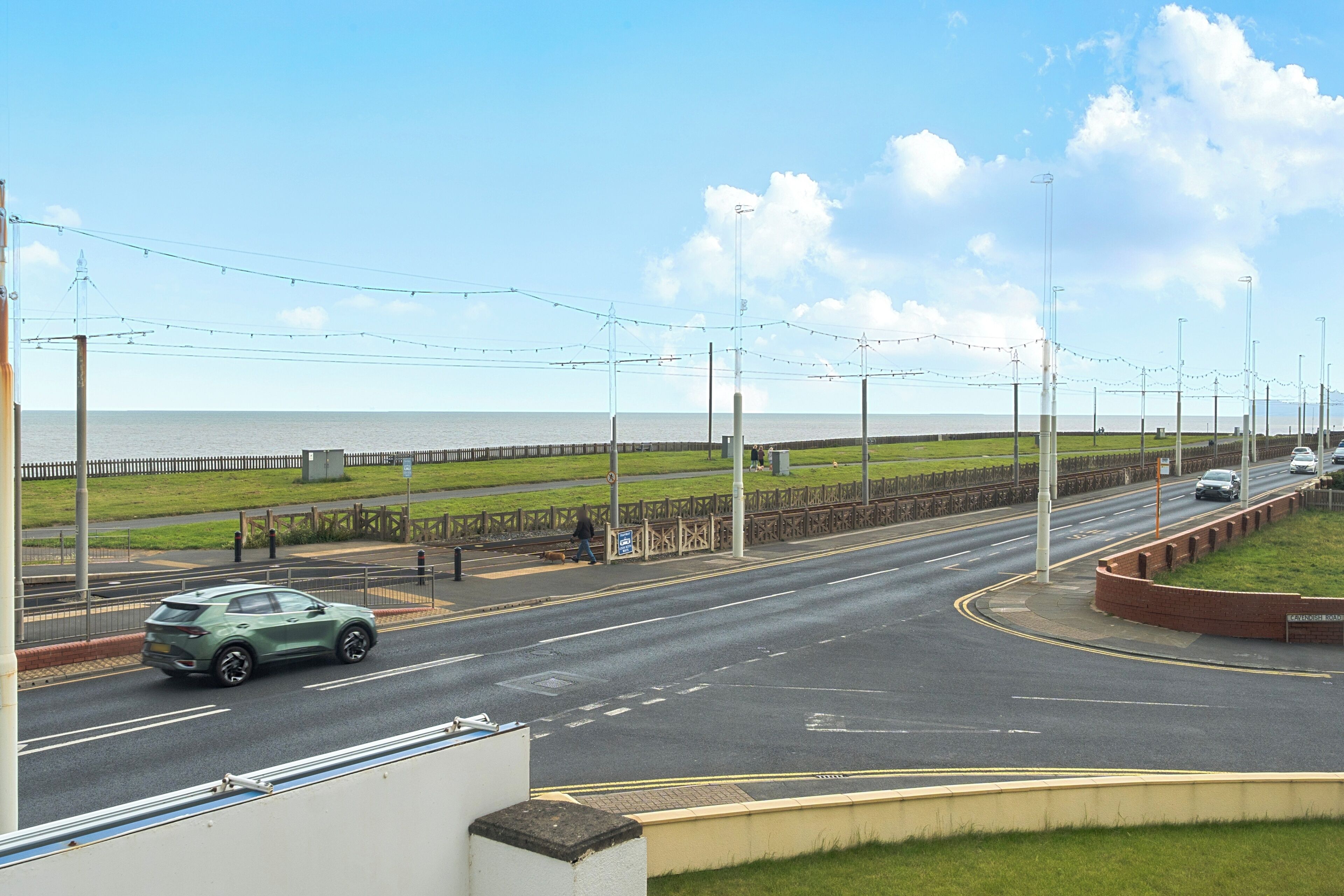 A road with a green car, a pedestrian crossing, and a grassy area with a fence.