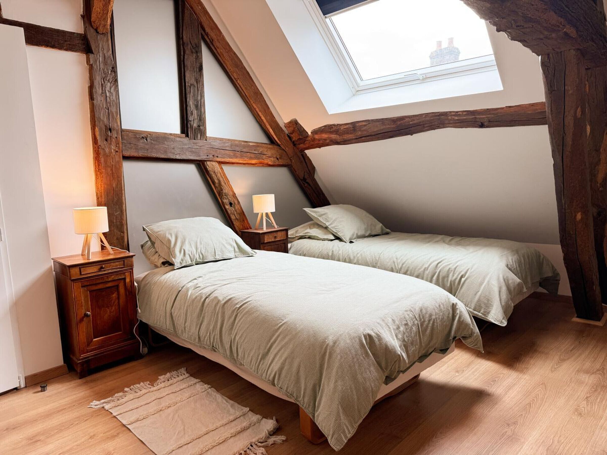 Une chambre à coucher aménagée dans un espace de type « loft », avec des poutres apparentes, deux lits, une lucarne et une table de chevet avec une lampe.