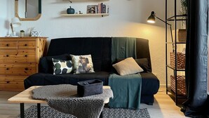 Ein gemütliches Wohnzimmer mit einem dunklen Sofa, einer Holzkommode, einem Spiegel, einem aufgehängten Hirschkopf, Regalen mit Büchern und Pflanzen und einer Lampe.