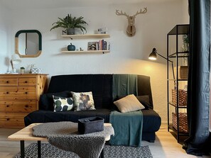 Ein gemütliches Wohnzimmer mit einem dunklen Sofa, einer Holzkommode, einem Spiegel, einem aufgehängten Hirschkopf, Regalen mit Büchern und Pflanzen und einer Lampe.