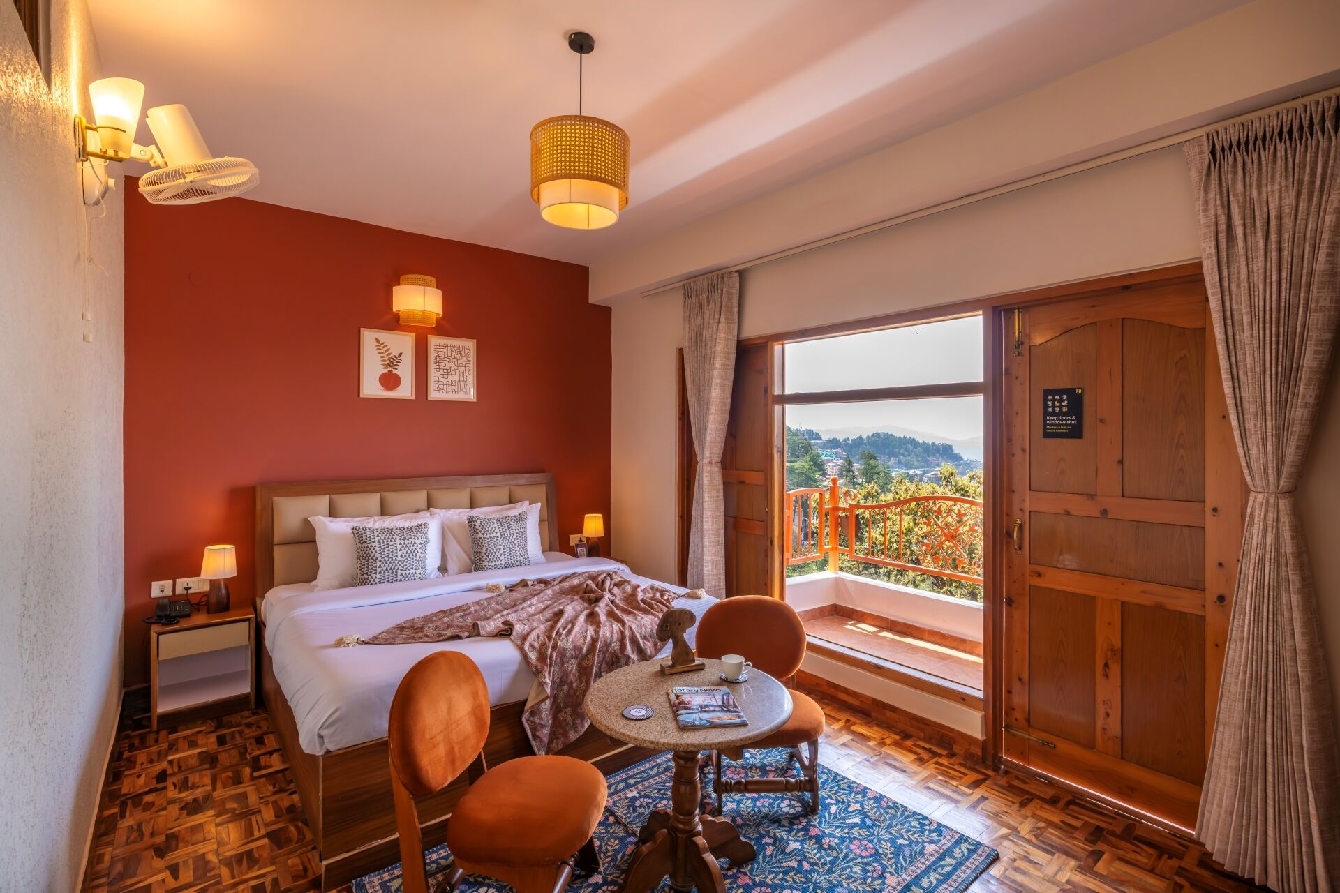 Habitación de hotel con cama, mesita de noche, lámpara, silla, mesa redonda y balcón con vistas.