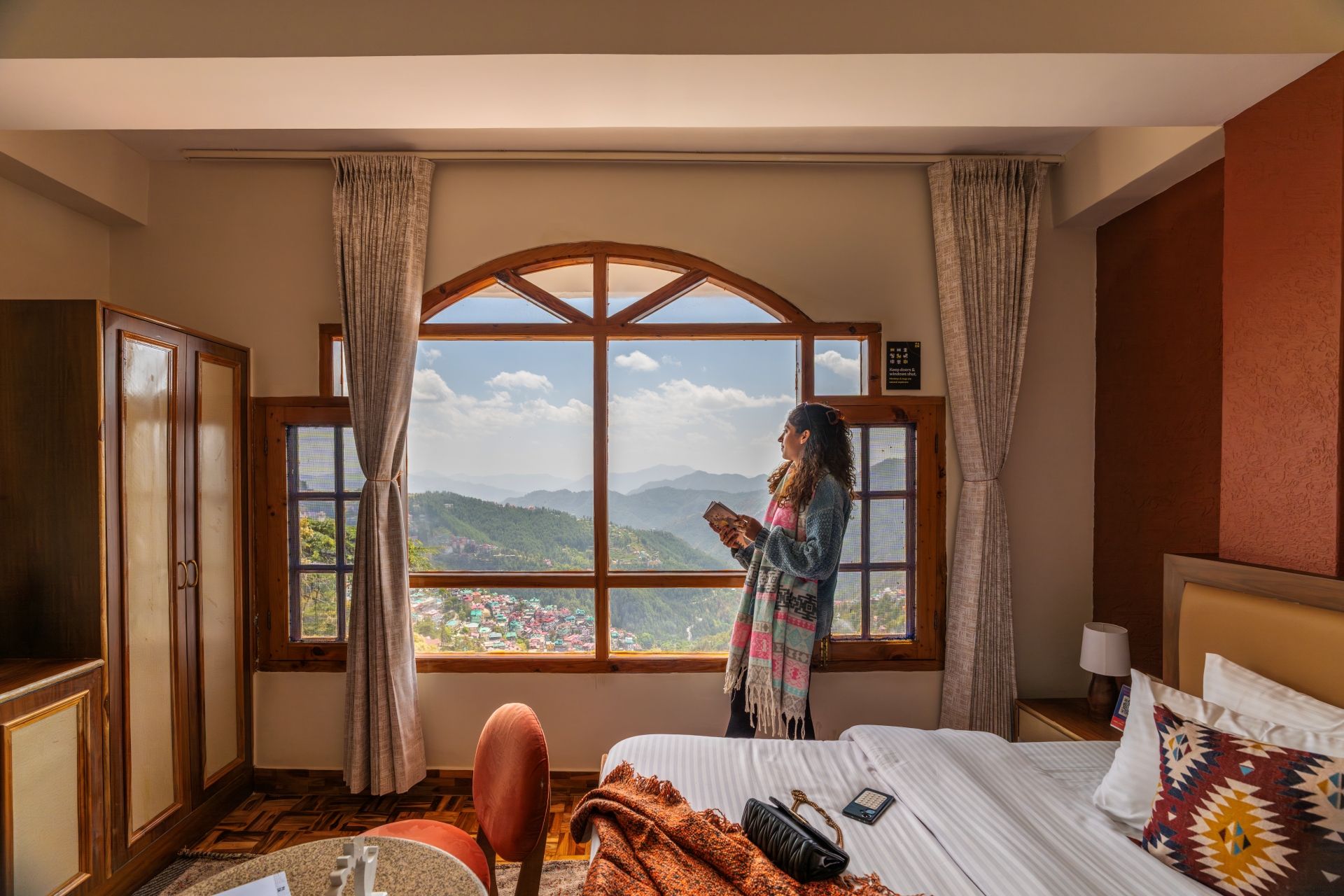 Habitación de hotel con un ventanal grande que ofrece vistas a la montaña, una cama con almohada estampada y un armario de madera.