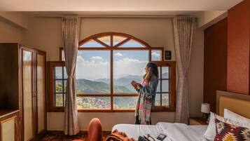 Habitación de hotel con un ventanal grande que ofrece vistas a la montaña, una cama con almohada estampada y un armario de madera.