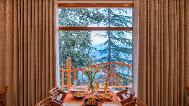 A dining table set with plates, bowls, and glasses, a window with curtains, and a view of trees and mountains. - PACK Shimla (Shimla)