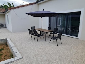 Eine Terrasse im Freien mit Tisch und Stühlen, einem dunklen Sonnenschirm und einer Glastür, die zu einem Innenbereich führt.