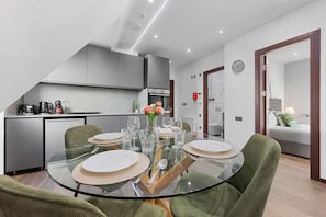 Eine moderne Küche mit Edelstahlgeräten, eine Essecke mit Glastisch und grünen Sitzmöbeln sowie ein Wohnbereich mit Sofa und Lampe.