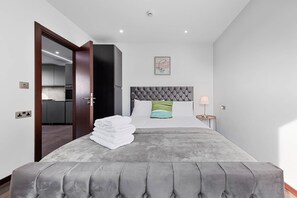 Ein Schlafzimmer mit einem Bett, einem Nachttisch, einer Lampe, einem gerahmten Bild an der Wand und Blick auf die Küche durch eine offene Tür.