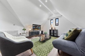 Ein modernes Schlafzimmer im Loftsstil mit schrägem Dach, großem Fenster, integriertem Fernseher, kleinem Beistelltisch und einer Couch.