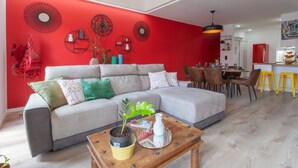 A modern living room with a red accent wall, a grey sofa, a wooden coffee table, and a dining area with yellow stools. - 4BR Duplex w Rooftop Views Workspaces (Lisbon)