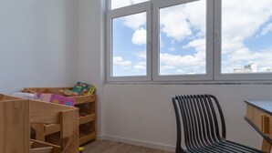 A room with a wooden playpen, a black chair, and a window with a city view. - 4BR Duplex w Rooftop Views Workspaces (Lisbon)