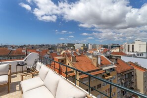 A rooftop terrace with a sofa, a small table, and a chair, overlooking a cityscape with red-tiled roofs and various buildings. - 4BR Duplex w Rooftop Views Workspaces (Lisbon)