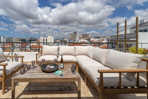 A rooftop terrace with a beige sectional sofa, a wooden coffee table, and a city view. - 4BR Duplex w Rooftop Views Workspaces (Lisbon)
