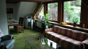 A living room with a sofa, a coffee table, a dining table with chairs, a kitchen area, and large windows with a view of greenery. - Vacation apartment panoramablick ( 85 m2 - 5 persons.) (Lennestadt)