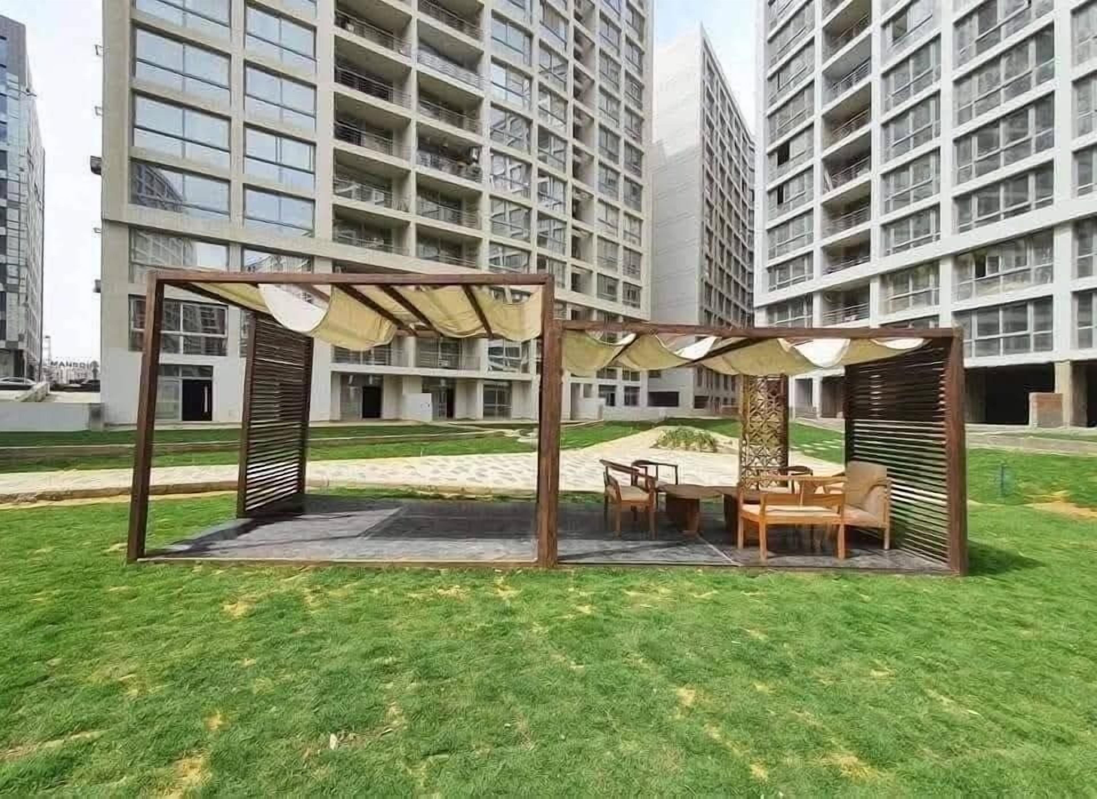 A modern outdoor seating area with a pergola structure, surrounded by well-maintained grass and a view of high-rise buildings.