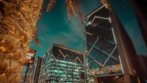 Un edificio moderno con fachada de vidrio y patrones geométricos, rodeado de palmeras y farolas.