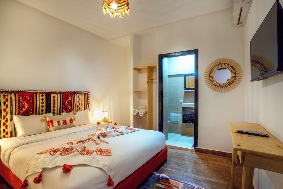 A hotel room with a bed, a TV, a wooden table, and a bathroom visible through an open door.