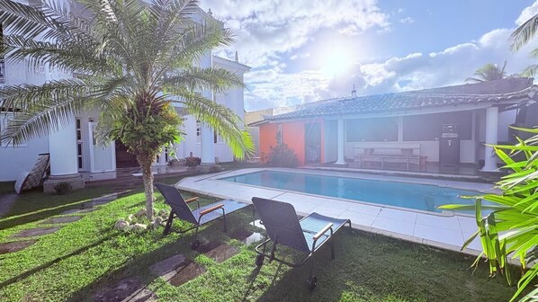 A swimming pool with lounge chairs, a palm tree, and a building with a tiled roof. - Terra do Sol Pousada (Porto Seguro)
