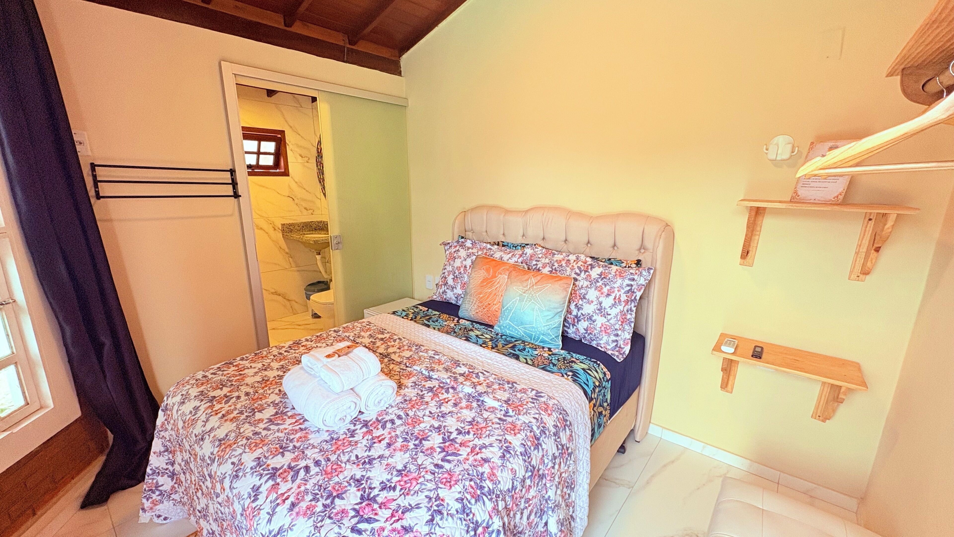A bedroom with a bed, a nightstand, a wooden shelf, and a bathroom visible through an open door.